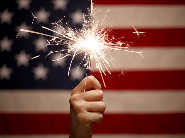 hand-holding-lit-sparkler-in-front-of-the-american-flag-for-4th
