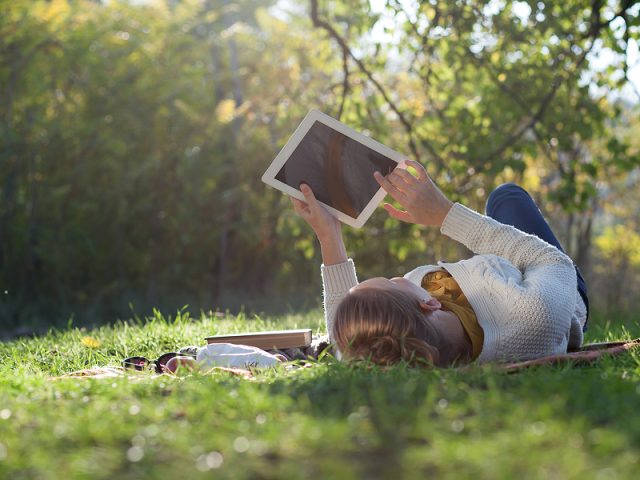 woman-lying-on-bedding-on-green-grass-with-ipad-during-picknic-i